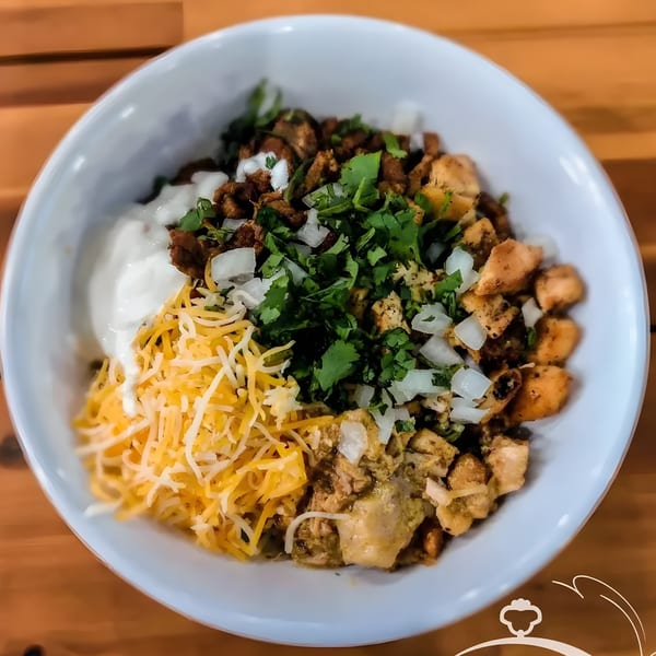 A Mexican bowl with meat, cheese, sour cream, cilantro, and onions on a wooden table.